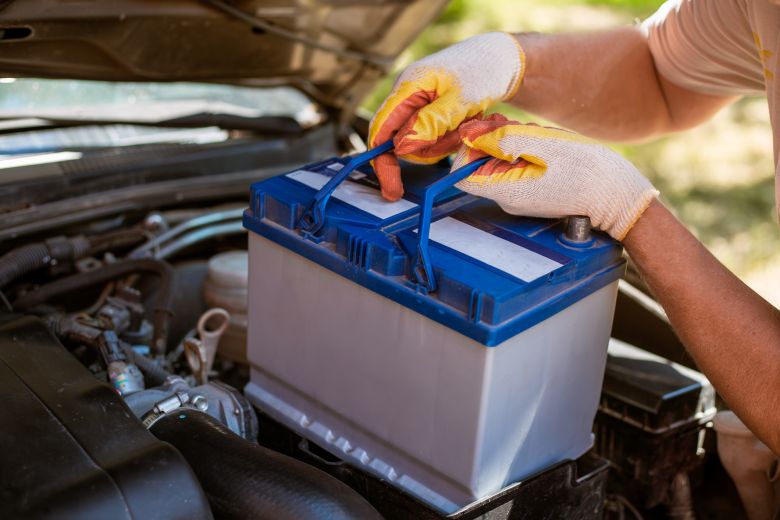 Person wearing gloves lifts a car battery with blue handles from an engine compartment of a vehicle with the hood open, working on car maintenance outdoors.
