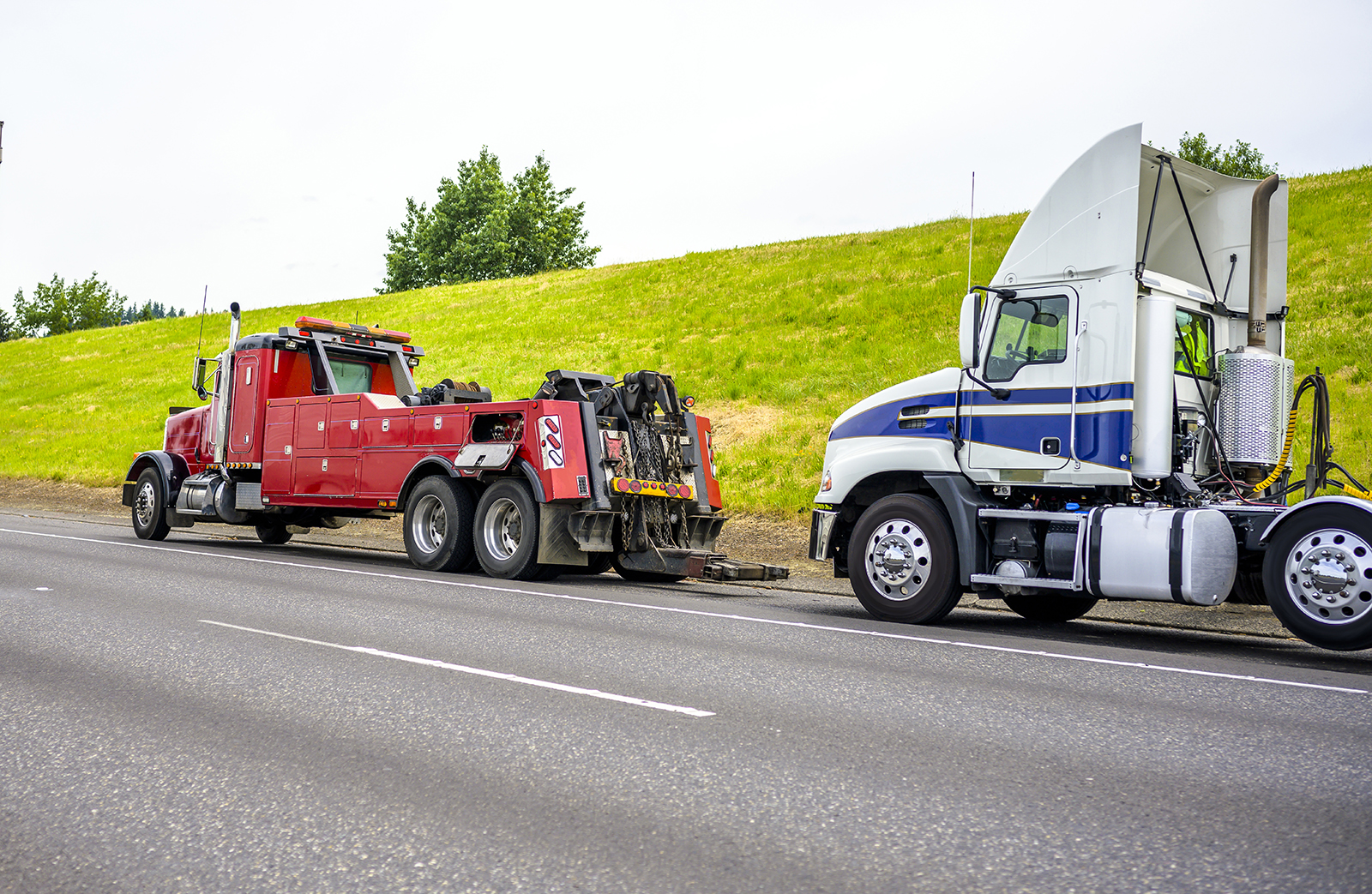 Powerful heavy-duty big rig mobile tow semi truck with emergency lights and towing equipment prepare to tow broken white big rig semi tractor standing out of service on the road side