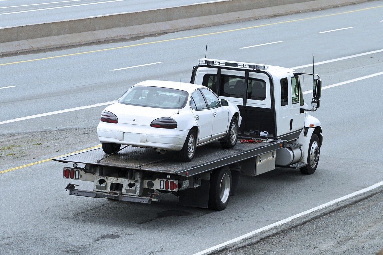 A white sedan is being transported on the flatbed of a tow truck driving down a multi-lane highway. The car appears to be undamaged, and the road is mostly empty.