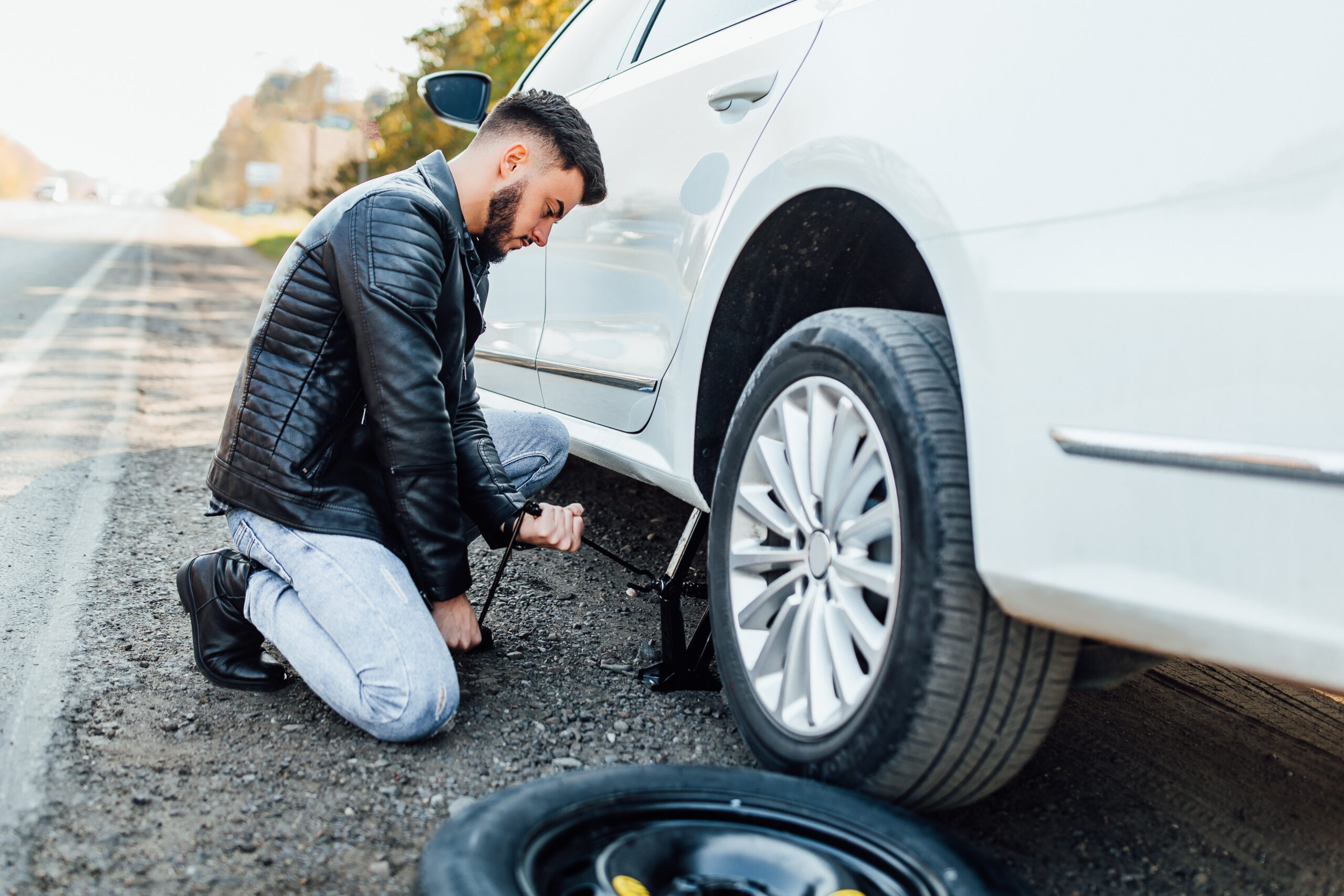 A man in a black leather jacket kneels beside a white car on the roadside, using a car jack to lift the vehicle. A spare tire lies on the ground in front of him.