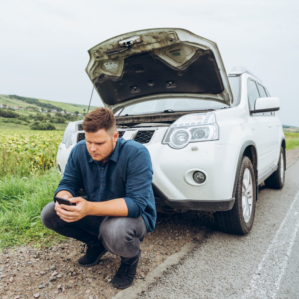 man sitting near broken car with phone, looking for help. road trip problems